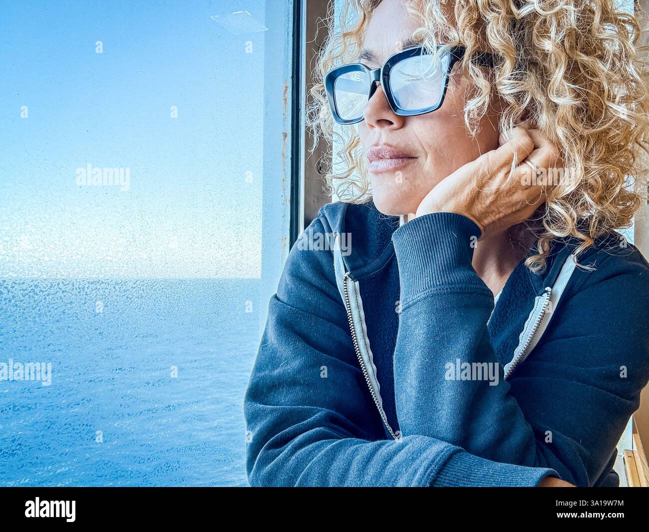 Schöne Frau mit Brille, die Freizeit mit Blick auf das Meer verbringt. Sie kümmert sich um sich, entspannt. Genießen Sie die Aussicht, schauen Sie durch das Fenster. Blonde und lockige Frauen mittleren Alters genießen die Aussicht. Ruhiger Morgen. Invia Commenti Stockfoto