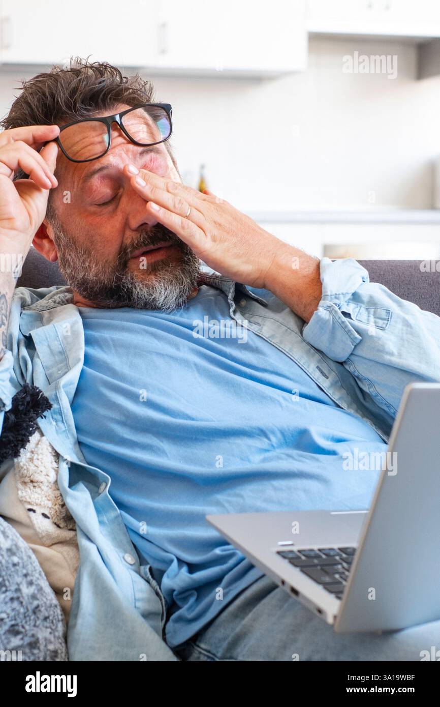 Ein müder Geschäftsmann zieht die Brille ab und spürt die Augenbelastung, wenn er online mit einem Laptop arbeitet, ein Kaukasier sitzt auf dem Sofa und reibt sich die Augen, fühlt Kopfschmerzen und Energielosigkeit Stockfoto