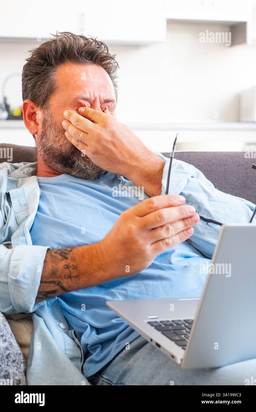 Ein müder Geschäftsmann zieht die Brille ab und spürt die Augenbelastung, wenn er online mit einem Laptop arbeitet, ein Kaukasier sitzt auf dem Sofa und reibt sich die Augen, fühlt Kopfschmerzen und Energielosigkeit Stockfoto