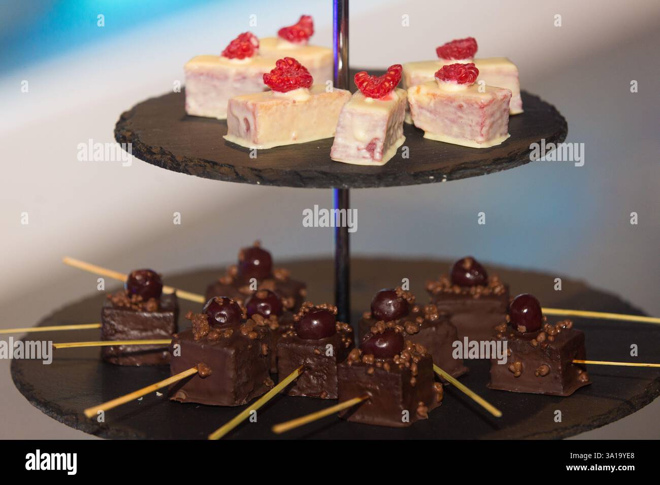 Dessertpräsentation bei einer Veranstaltung. Miniaturkuchen (Himbeere aus weißer Schokolade und Kirsche aus dunkler Schokolade) auf einem gestuften Schieferständer Stockfoto