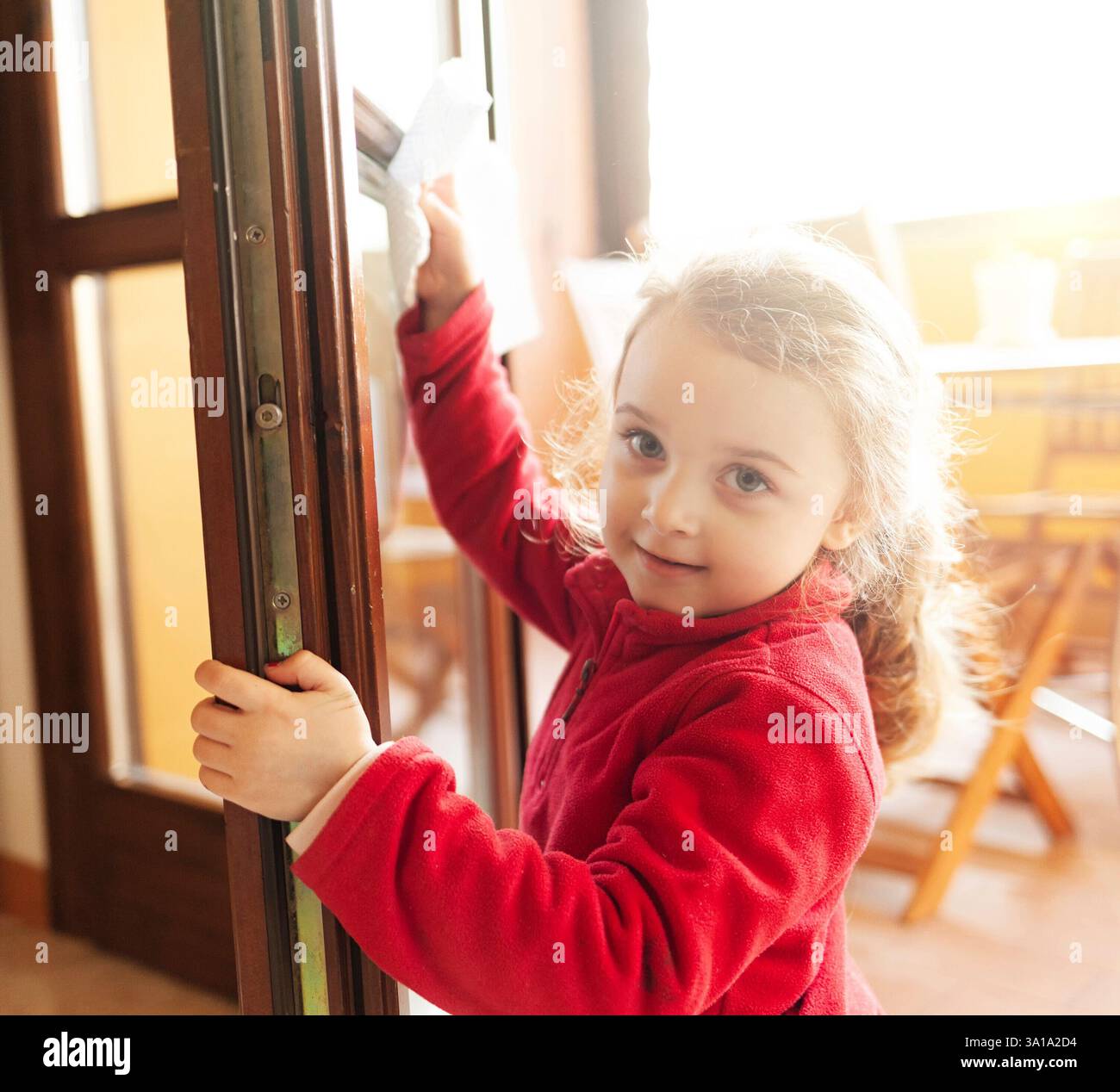Das dreijährige Mädchen hilft bei der Haushaltsreinigung. Sie reinigt die Fenster mit einem Blatt Papier. Idee, die Zeit während der Kovid-19-Coronavirus-Quarantänezeit zu verstreichen. Stockfoto