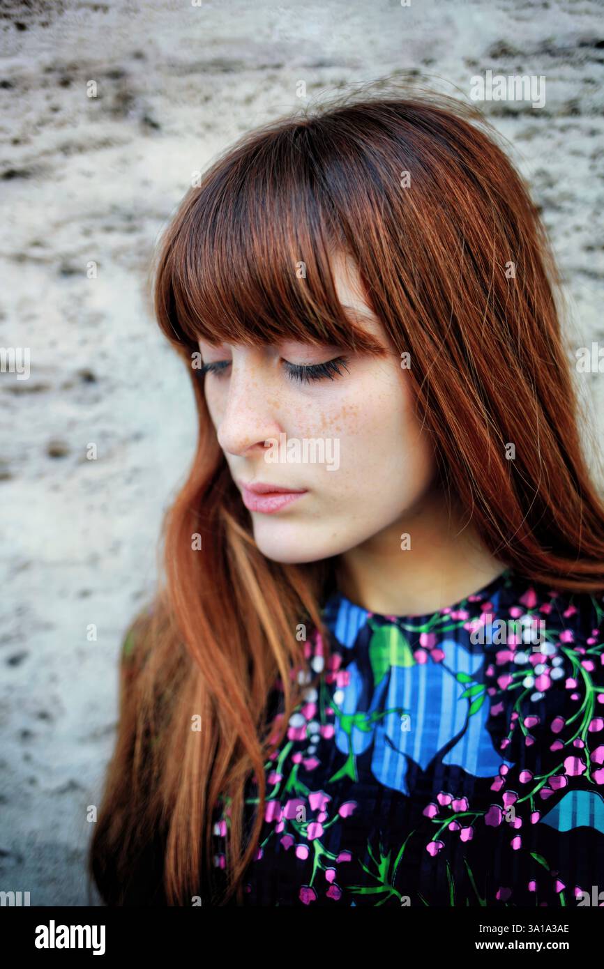 Eine junge Frau mit langen braunen Haaren und Sommersprossen, die ein Oberteil mit Blumenmuster trägt, blickt mit einem nachdenklichen Ausdruck nach unten Stockfoto
