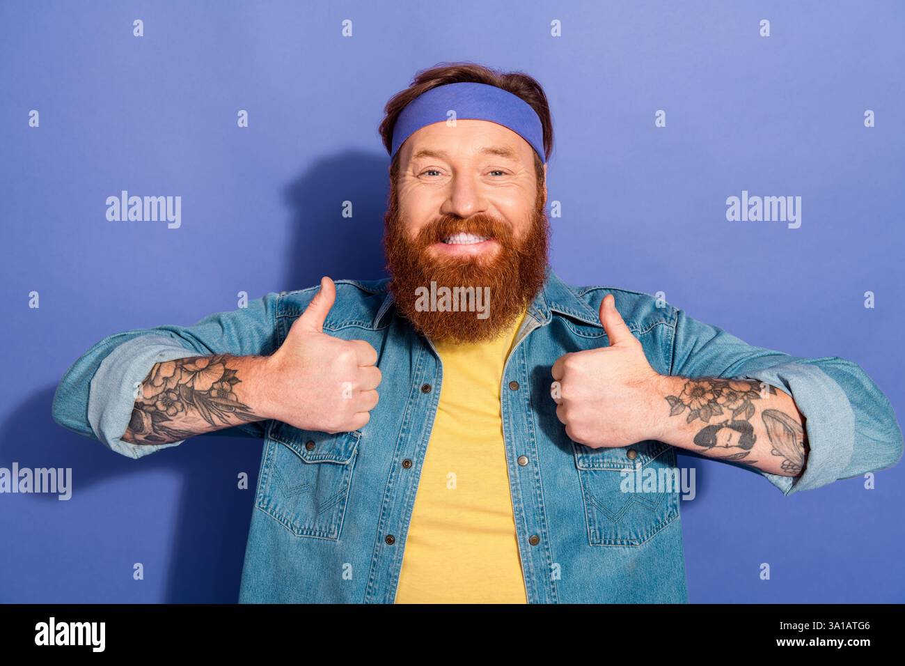 Fröhlicher bärtiger Mann mit Tätowierungen, die die Daumen in lässigem Stil vor violettem Hintergrund hochlegen und Retro- und Hipster-Ästhetik verkörpern. Stockfoto