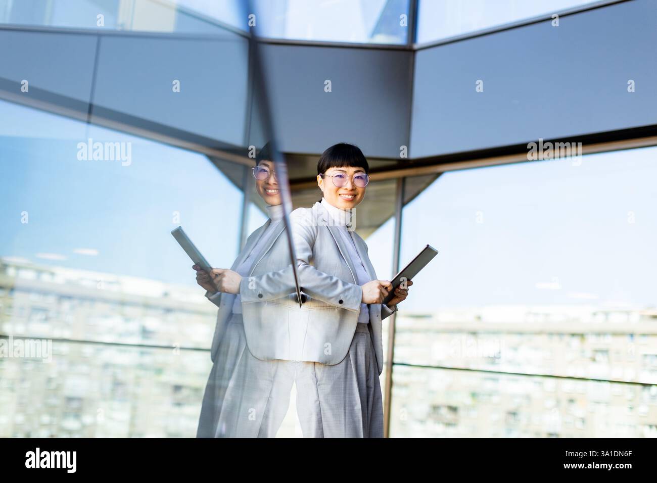 Ein selbstbewusster Unternehmer steht in einem hellen, modernen Büro und hält ein Tablet. Die atemberaubende Skyline der Stadt ist durch große Glasfenster zu sehen. Stockfoto