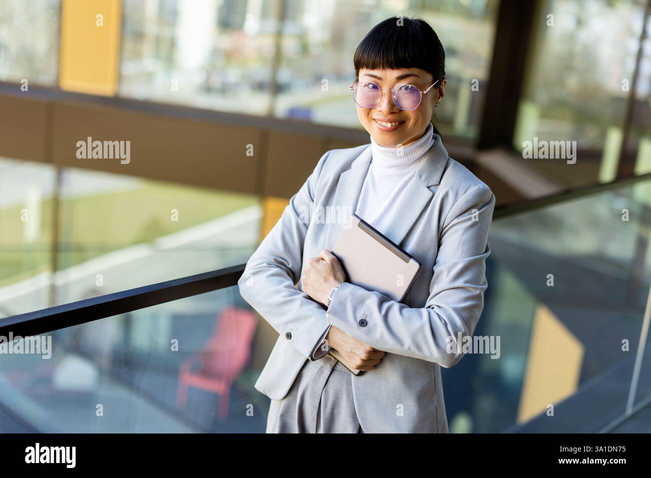 Asiatische Frau steht in einer eleganten Büroumgebung und hält ein Tablet unter dem Arm. Sie trägt einen maßgeschneiderten grauen Anzug und eine Brille und strahlt Selbstvertrauen aus wie Sch Stockfoto