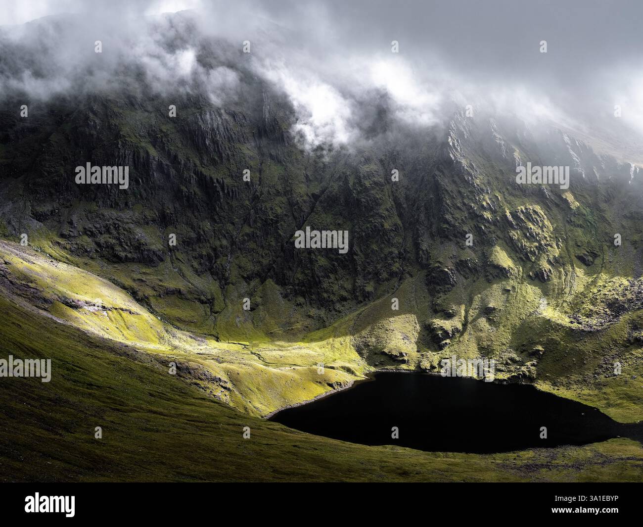 Ein atemberaubender Blick auf zerklüftete Berge, die einen ruhigen dunklen See in Irland umgeben. Die Landschaft ist lebendig mit moosigem Grün und versteckten Schatten Stockfoto