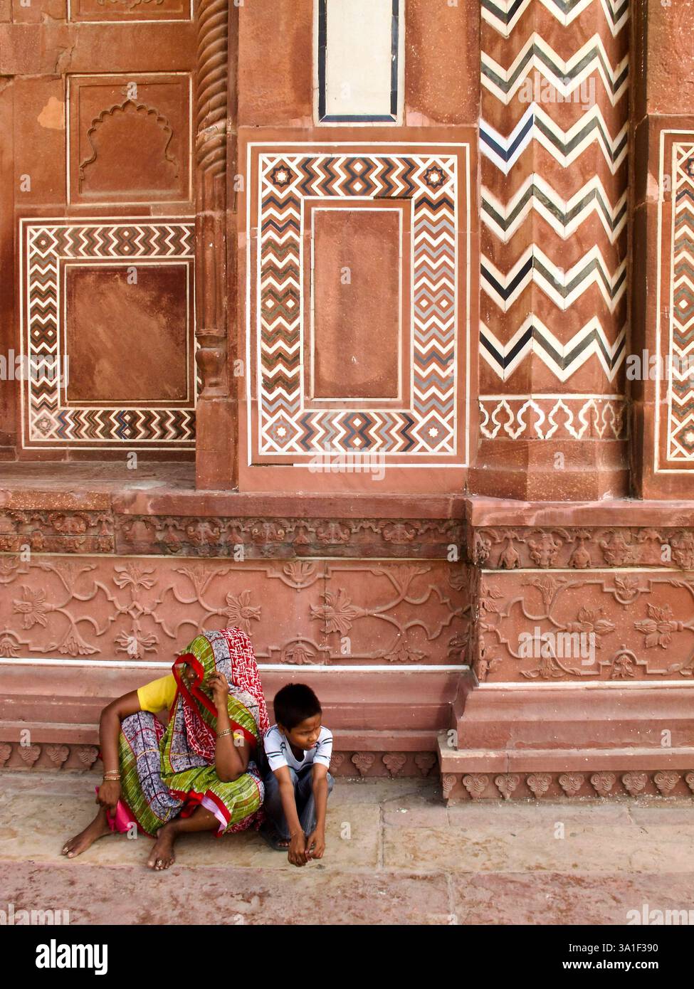 Frau und Kind ruhen an einer roten Sandsteinwand mit geometrischer Intarsie im Sirhi Darwaza, Taj Mahal Komplex, Agra, Indien. Stockfoto