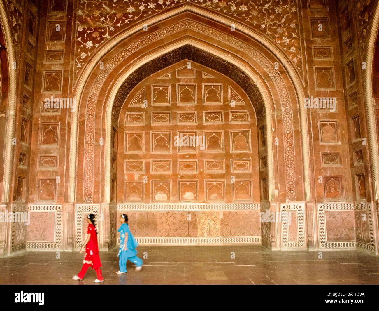 Zwei Frauen gehen an einem geschnitzten roten Sandsteinbogen mit floralen und geometrischen Details in der Moschee des Taj Mahal Komplexes in Agra, Indien vorbei. Stockfoto