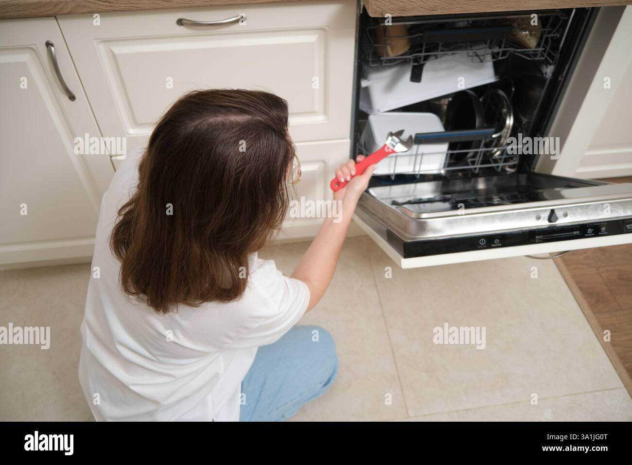 Frau, die einen Schraubenschlüssel benutzt, um einen offenen Geschirrspüler in einer modernen Küche zu reparieren. Haushaltsgerätewartung und Hausreparaturkonzept. Stockfoto