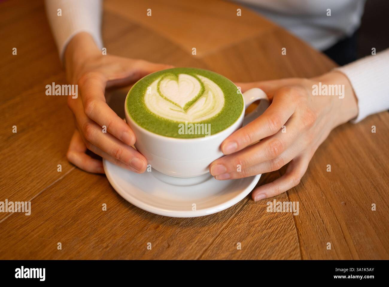 Frauenhände halten eine weiße Tasse Matcha Latte, verziert mit einer herzförmigen Latte Art, die auf einem Holztisch platziert wird, und schaffen eine gemütliche und einladende Atmosphäre Stockfoto