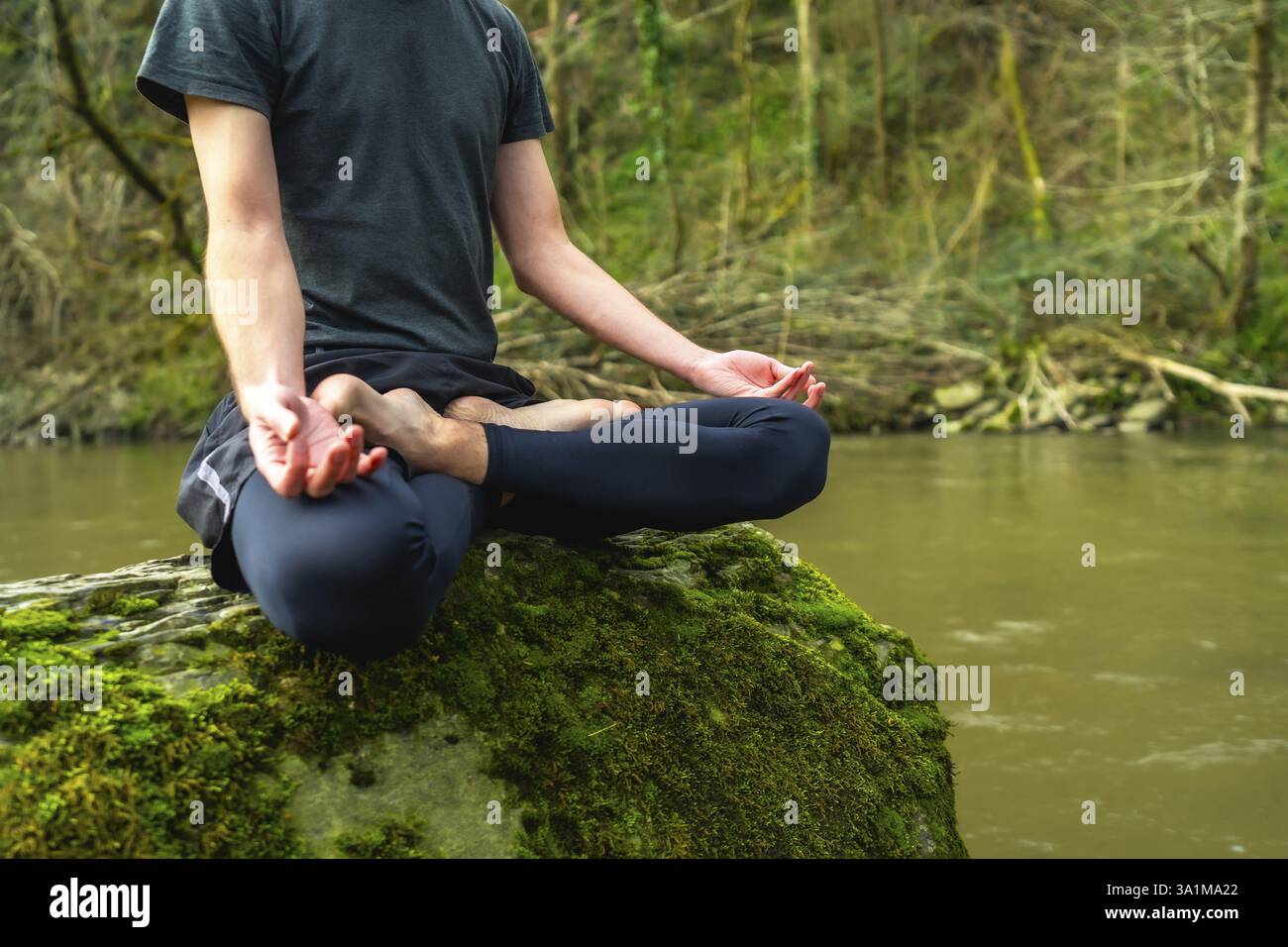 Junger Mann, der in Lotusposition auf moosigem Felsen am Fluss meditiert, friedliche Natur genießt und inneren Frieden findet Stockfoto