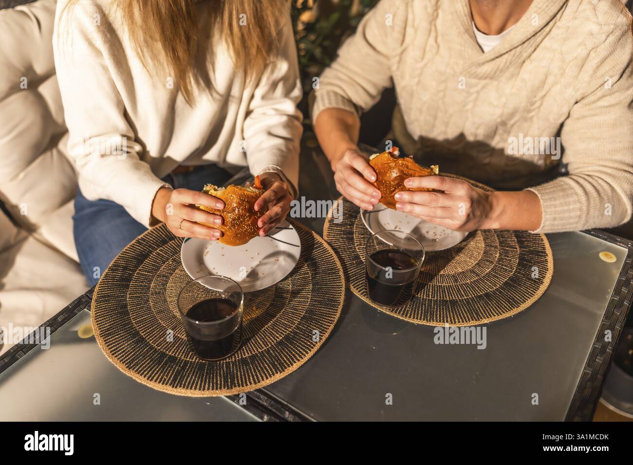 Ein Paar, das einen Moment beim Essen von Burgern und Trinken, wahrscheinlich Wein oder Limonade, mit Tischsets an einem Tisch sitzt Stockfoto