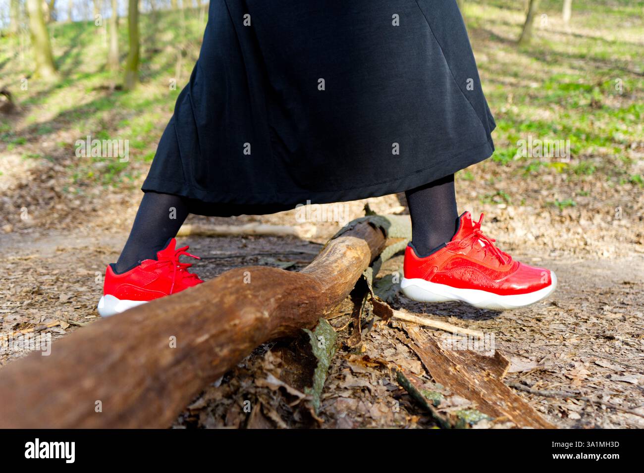 Eine Person in schwarzer Kleidung mit roten Turnschuhen schlendert durch einen üppigen Wald und geht über einen gefallenen Ast auf einem unbefestigten Weg, umgeben von Grün Stockfoto