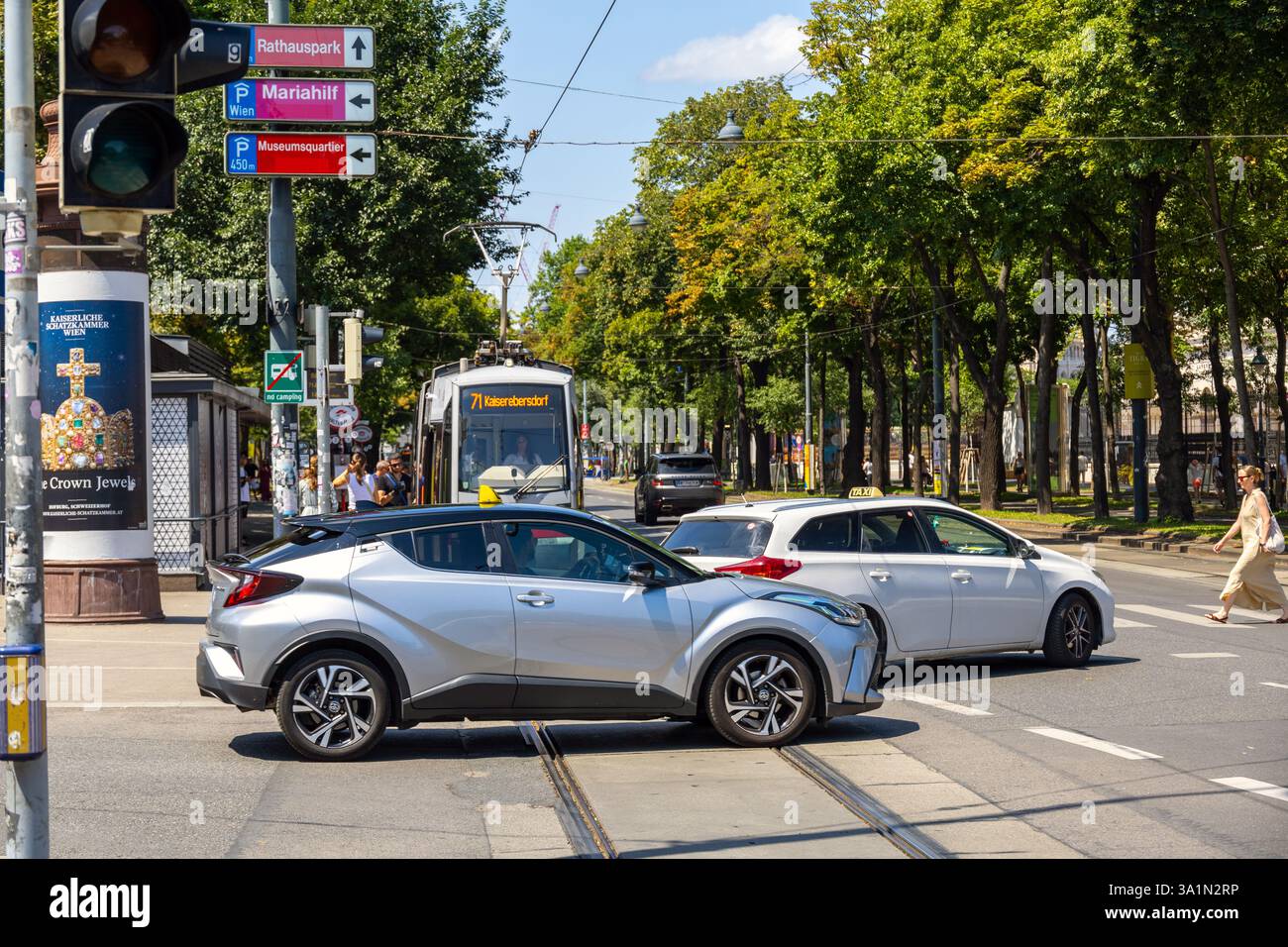 Sommerverkehr in Wien mit silbernem Toyota C-HR und weißem Toyota Auris-Taxi an einer Straßenbahnüberfahrt in der Nähe von grünem Park und Straßenschildern Stockfoto