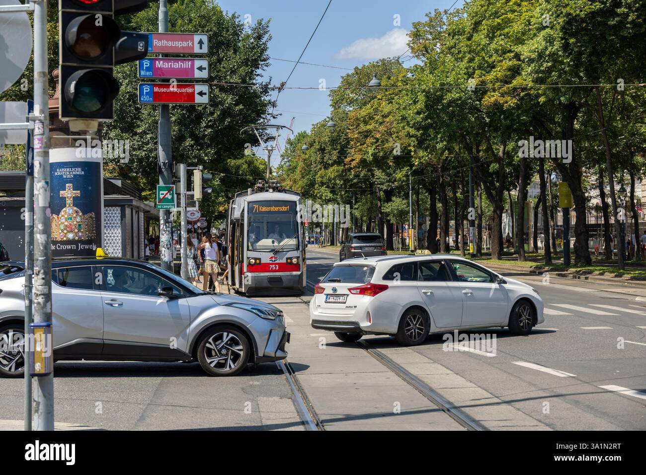 Sommerverkehr in Wien mit silbernem Toyota C-HR und weißem Toyota Auris-Taxi an einer Straßenbahnüberfahrt in der Nähe von grünem Park und Straßenschildern Stockfoto