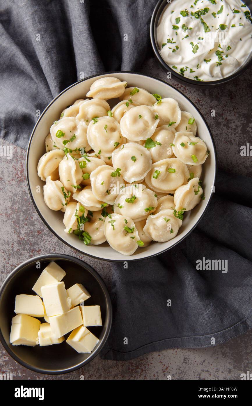 Russische Pelmeni-Knödel mit Sauerrahm, Butter und Kräutern aus nächster Nähe auf dem Tisch. Vertikale Draufsicht von oben Stockfoto