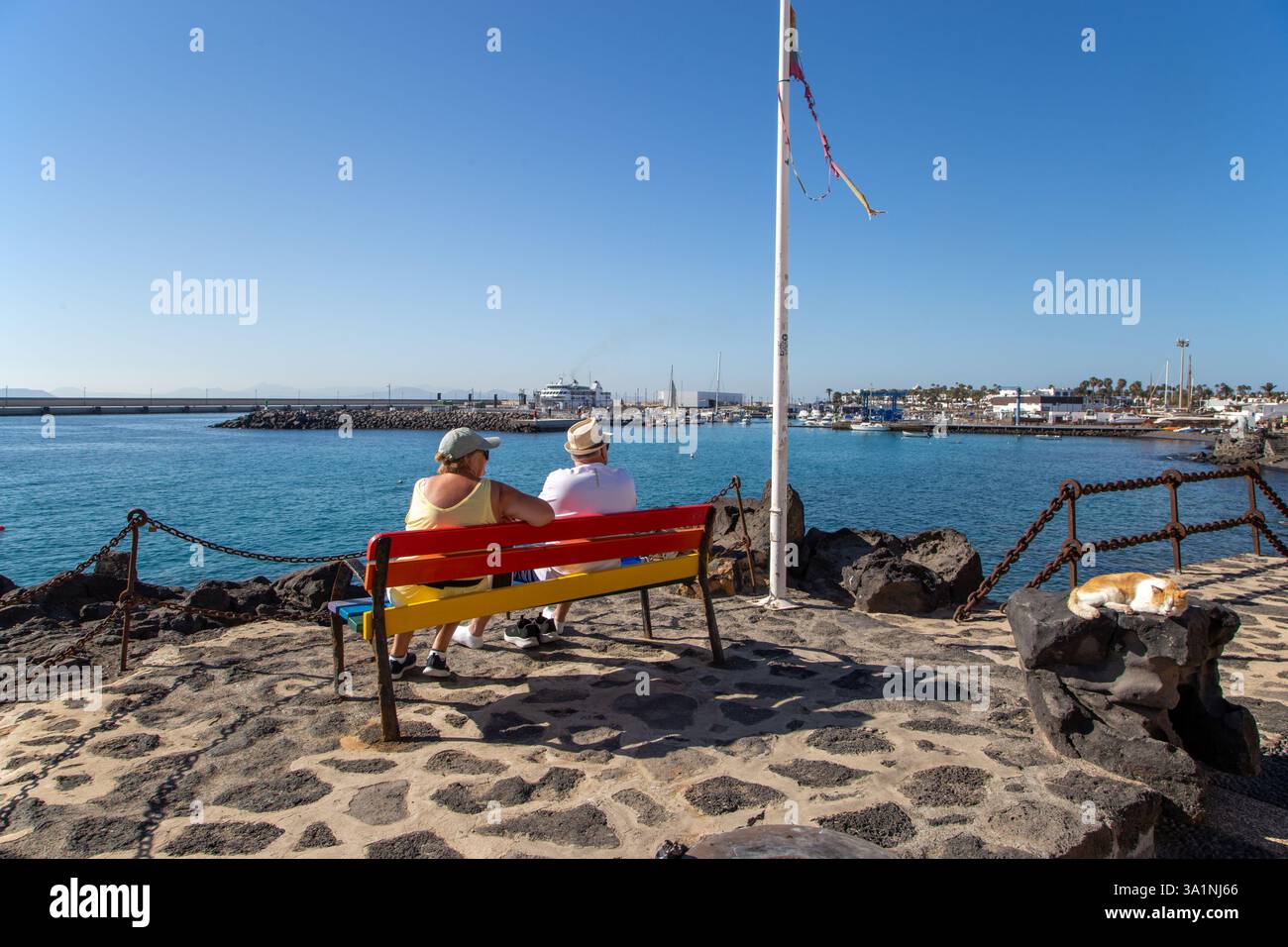Pärchen saßen auf einer Bank mit Blick auf den Hafen im Kanarischen Resort Playa Blanca Lanzarote Spanien mit einer Katze, die auf den Felsen schlief Stockfoto