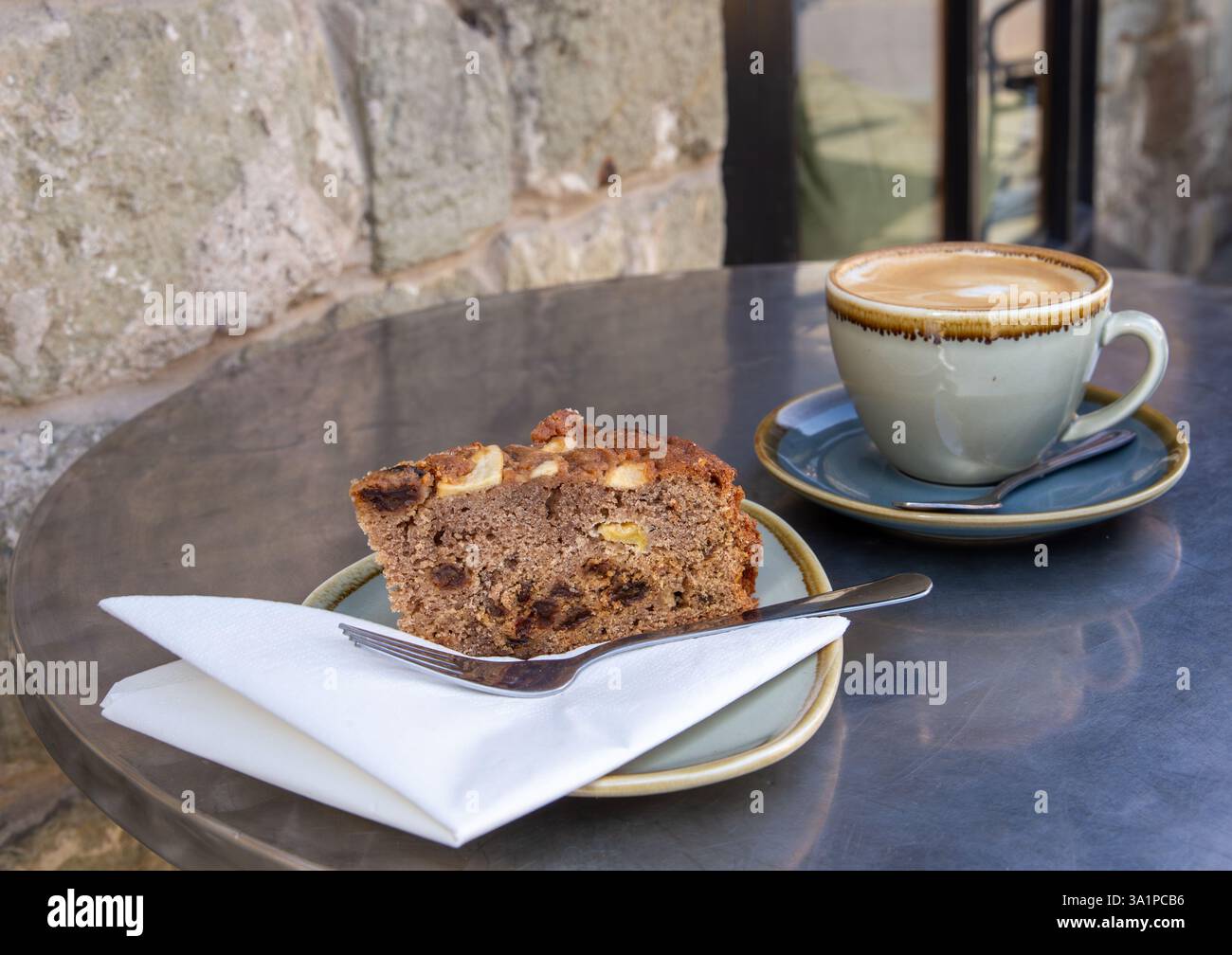 Nahaufnahme eines Stücks Dorset Apfelkuchen und Cappuccino auf einem Tisch mit Steinmauern in einem Café im Freien Stockfoto