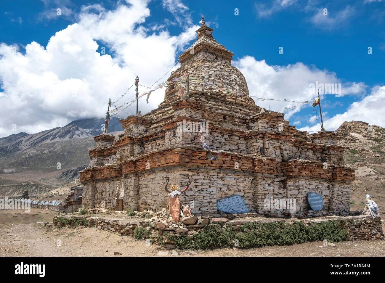 Heilige Steine, die mit Mantras beschriftet sind, bilden eine Mani-Mauer in der Nähe von Nar, Nepal, die alte Stupas und zerklüftete Gipfel unter einem dramatischen Himmel zeigt. Stockfoto