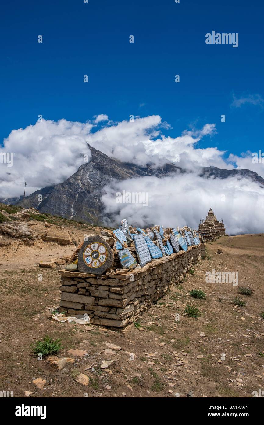 Heilige Steine, die mit Mantras beschriftet sind, bilden eine Mani-Mauer in der Nähe von Nar, Nepal, die alte Stupas und zerklüftete Gipfel unter einem dramatischen Himmel zeigt. Stockfoto