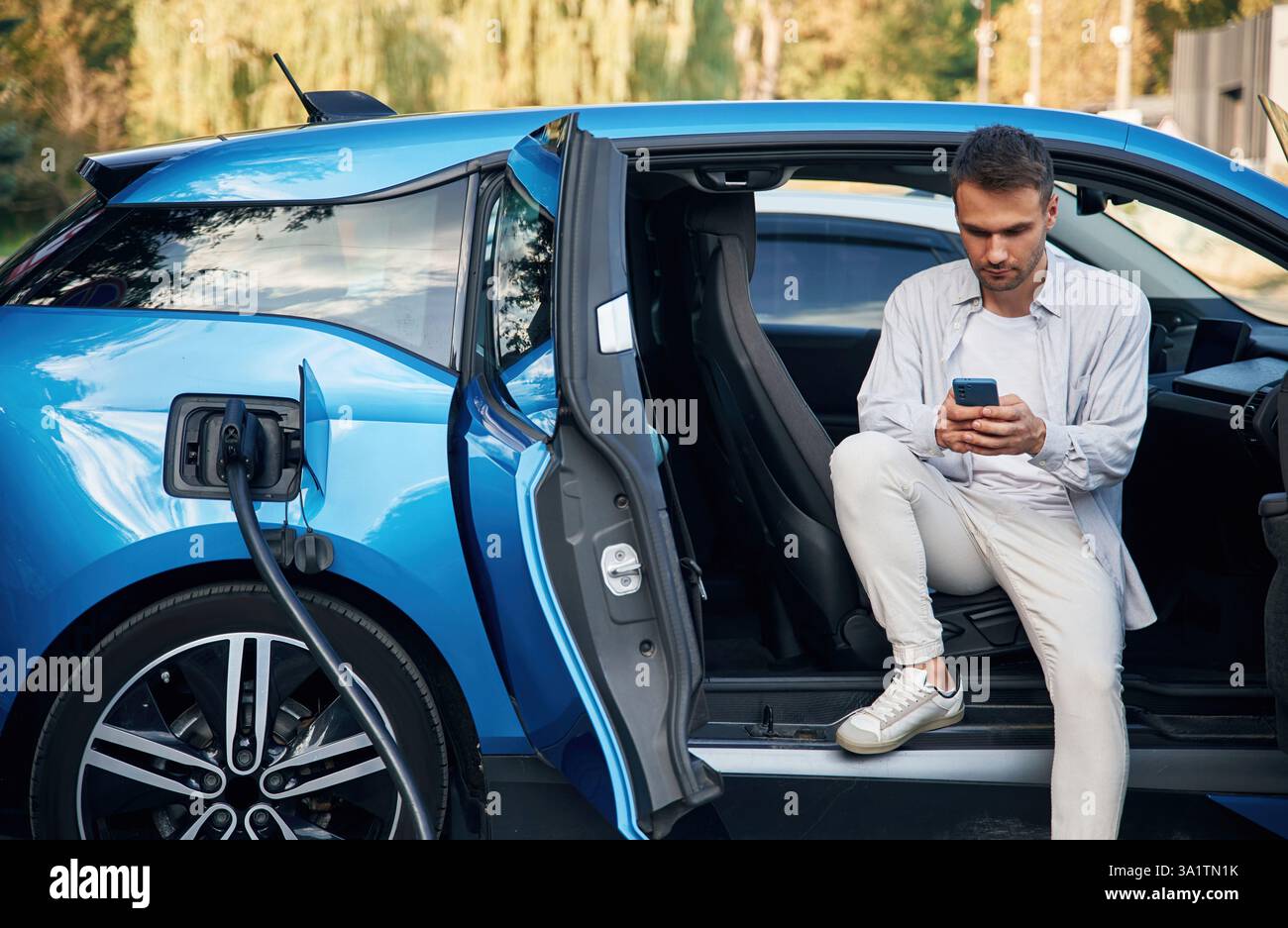 Schöne Person mit Smartphone. Mann mit blauem Elektroauto auf der Ladestation. Stockfoto