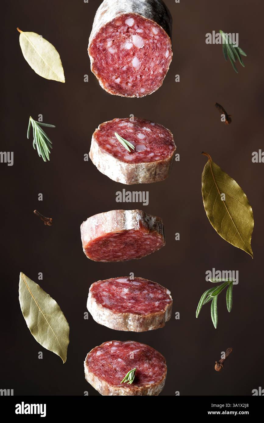 Salamifleischscheiben, die in der Luft hängen und von herabfallenden Kräutern und Gewürzen umgeben sind. Stockfoto