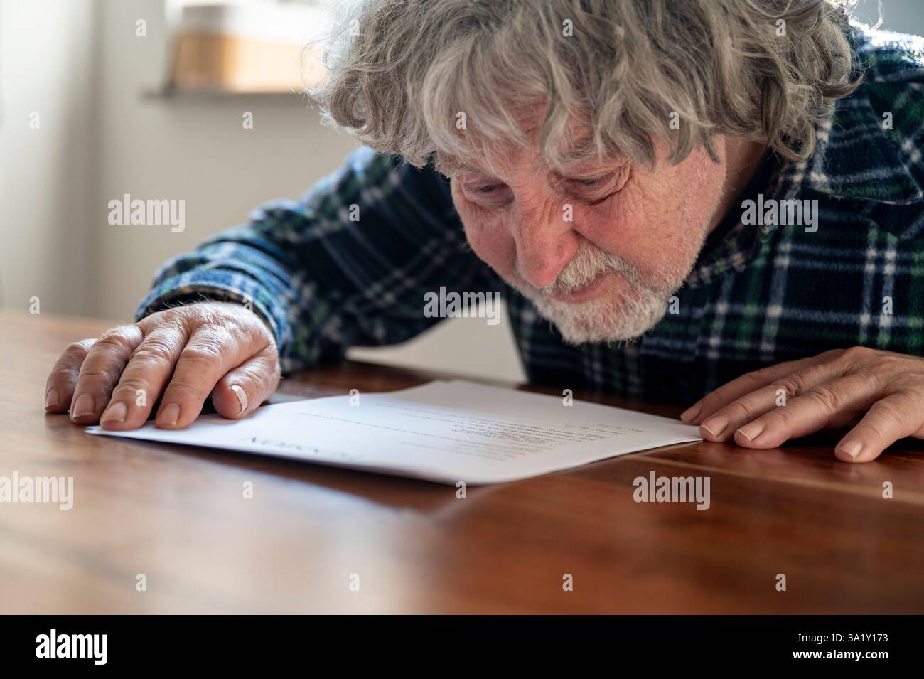 Ein älterer Mann mit grauen Haaren und Bart beugt sich dicht vor, um ein Dokument auf einem Holztisch zu lesen, das konzentriert und konzentriert wirkt. Stockfoto