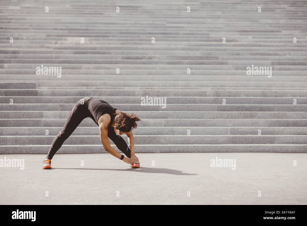 Dehnung nach dem Training. Afroamerikanische Frau mit Fitness-Tracker und kabellosen Kopfhörern macht Übungen für Beine Stockfoto