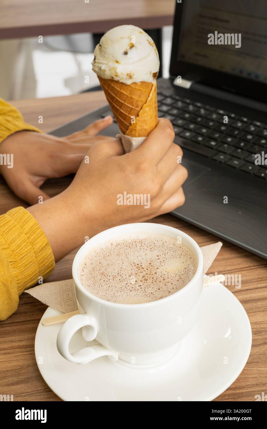 Ein Mädchen genießt einen süßen Moment Eis und heiße Schokolade, während es ihre Laptop-Aufgaben erledigt und mühelos Arbeit und köstliche tr in Einklang bringt Stockfoto
