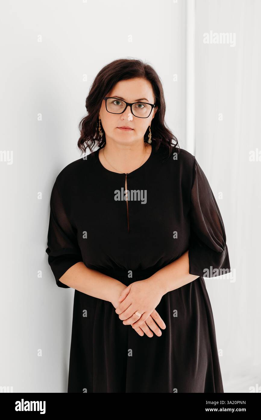 Studio-Porträt einer selbstbewussten 40-jährigen Frau, die ein elegantes schwarzes Outfit und eine Brille vor einem sauberen weißen Hintergrund trägt. Stockfoto