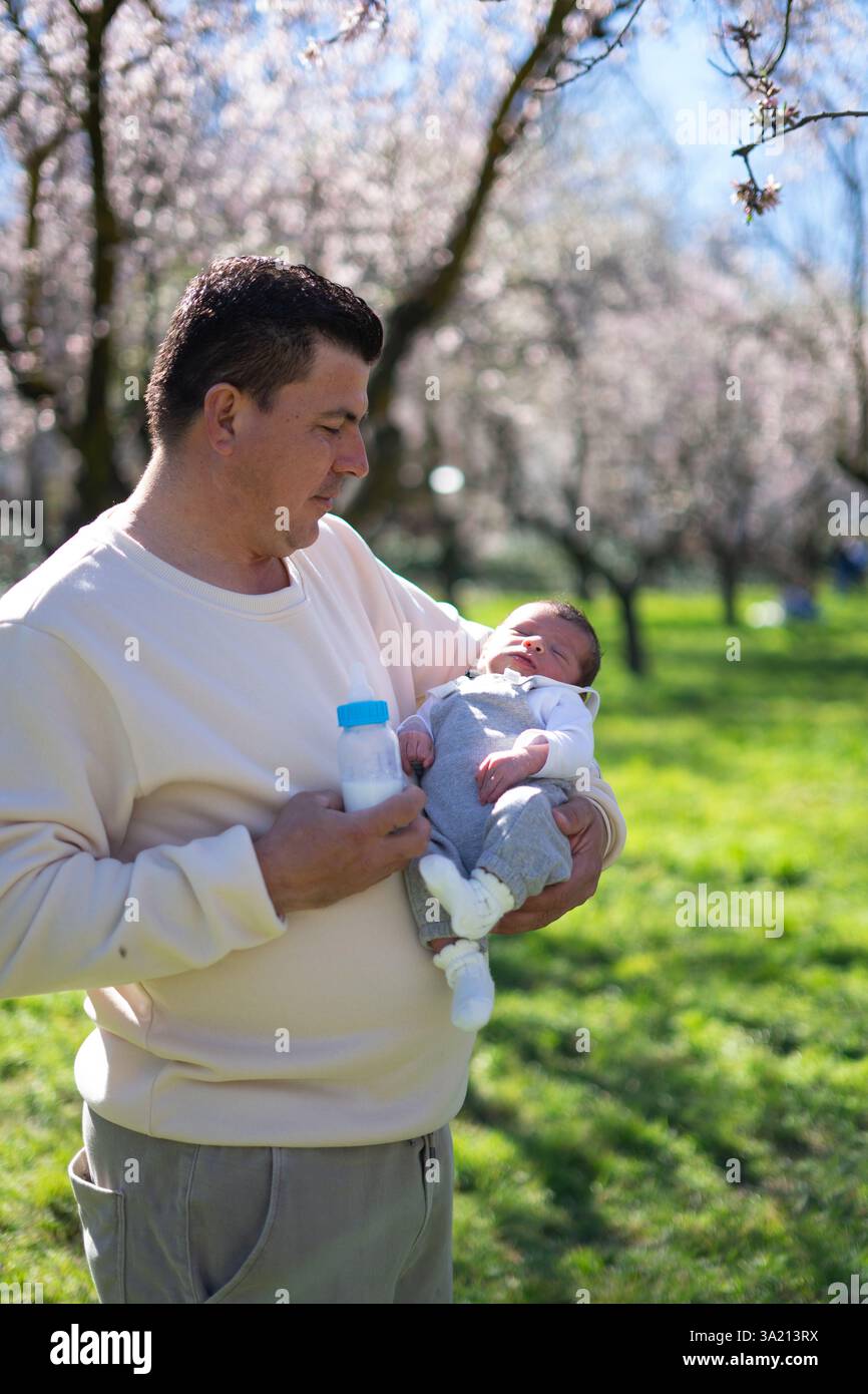 Vater, der sein neugeborenes Baby in der Hand hält, während er eine Milchflasche in einem wunderschönen blühenden Obstgarten hält, und einen friedlichen Moment zusammen genießt Stockfoto