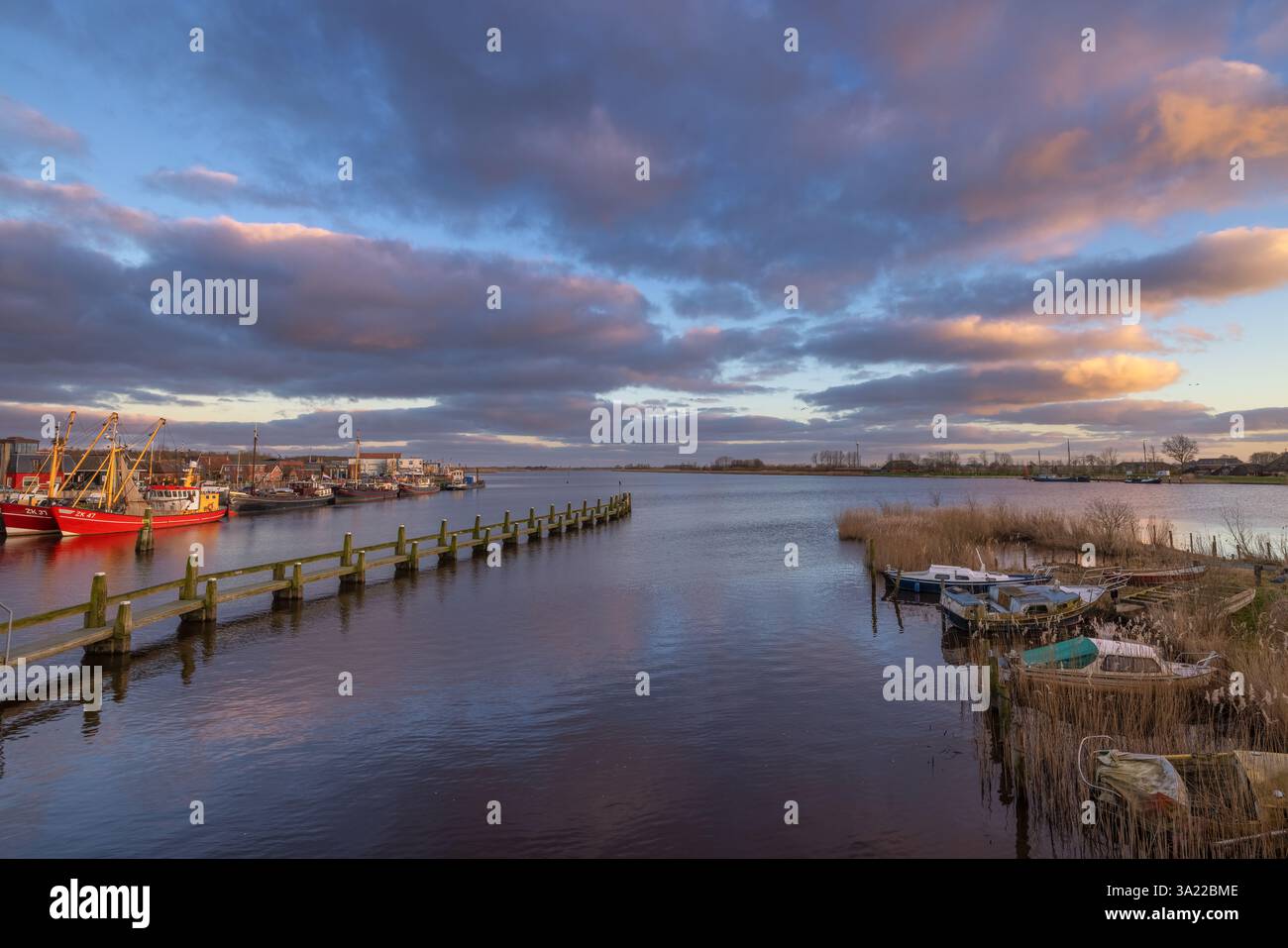 Sonnenuntergang über dem kleinen Dorf Zoutkamp in den Niederlanden mit Reflexionen des Himmels im Kanal und Fischereifahrzeugen im Hintergrund. Stockfoto