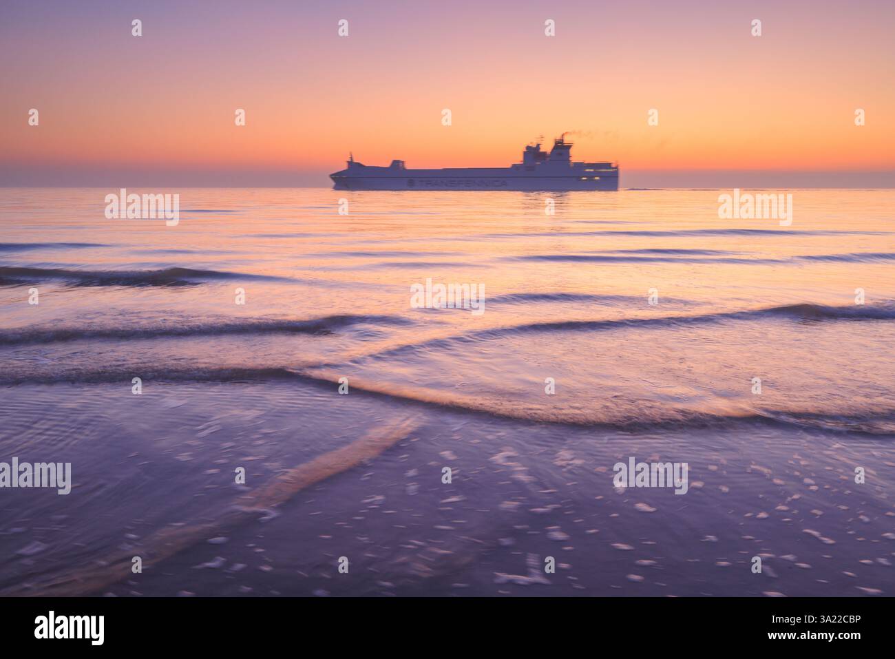 Ein großes Schiff segelt am Horizont bei Sonnenuntergang über der Nordsee mit einem farbenfrohen klaren Himmel und Wellen im Vordergrund vorbei. Stockfoto