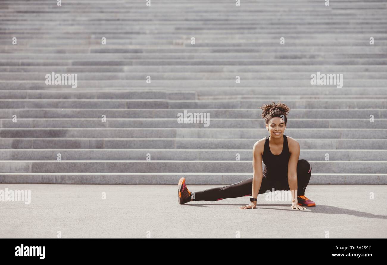 Erschwinglicher Sport im Freien. Lächelnde afroamerikanische Frau in Sportkleidung mit Fitness-Tracker und Kopfhörer tun Stretch für Bein, in der Nähe der Stadt Treppe Stockfoto