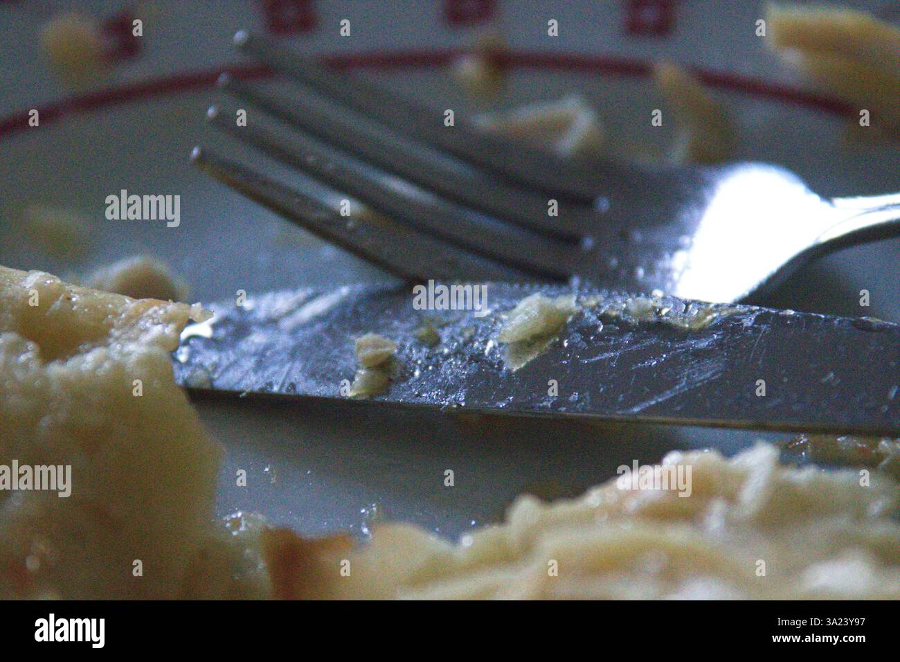 Ein genauer Blick auf eine Gabel und ein Messer, die auf einem Teller liegen, auf dem Reste von Lebensmitteln aufbewahrt werden, was auf eine kürzliche Mahlzeit hinweist. Die Einstellung ist schwach beleuchtet, wodurch eine Invit entsteht Stockfoto