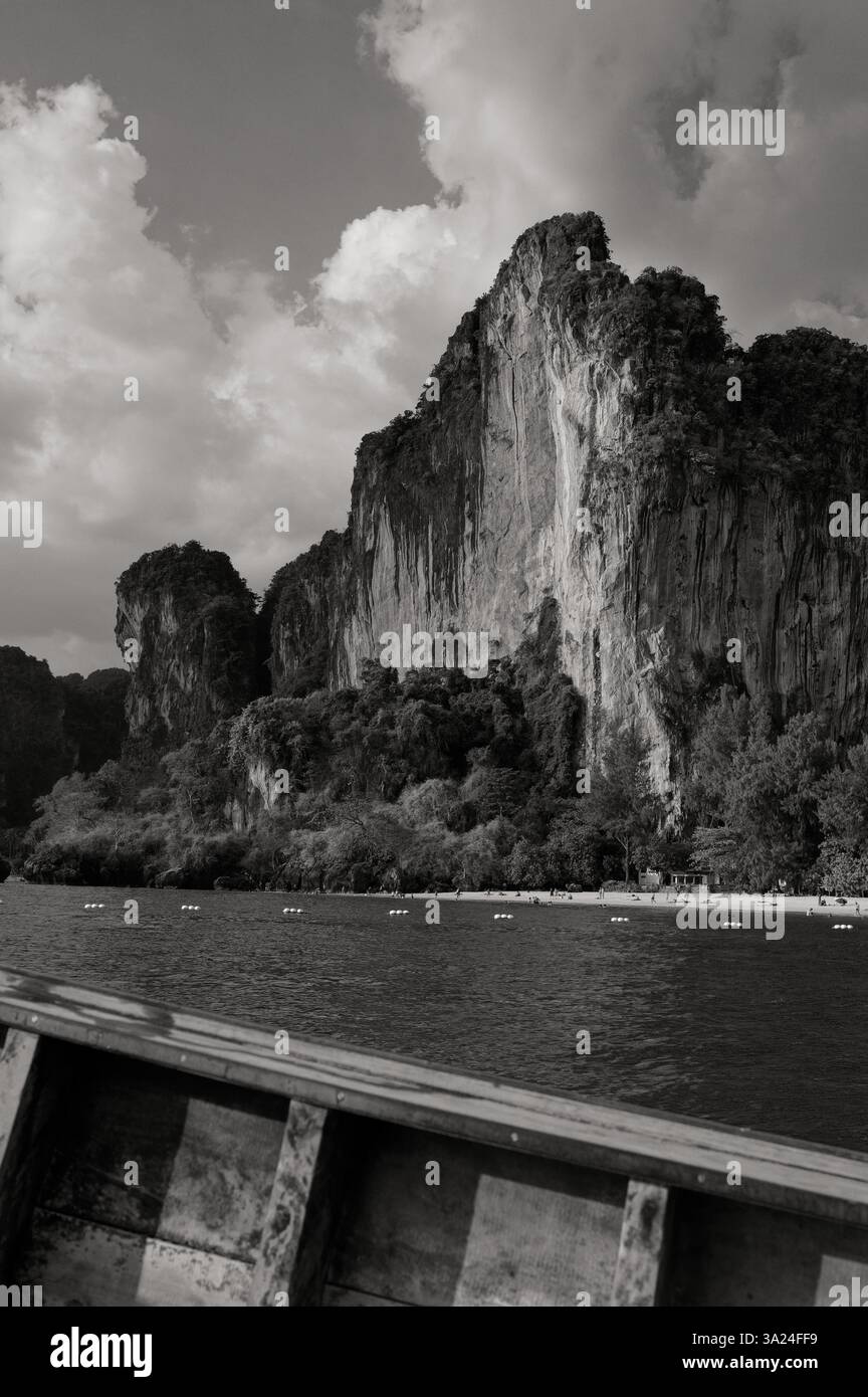 Dramatische Kalksteinklippen überragen einen abgeschiedenen Strand in Railay, gefangen in S&W von einem traditionellen Holzboot Stockfoto