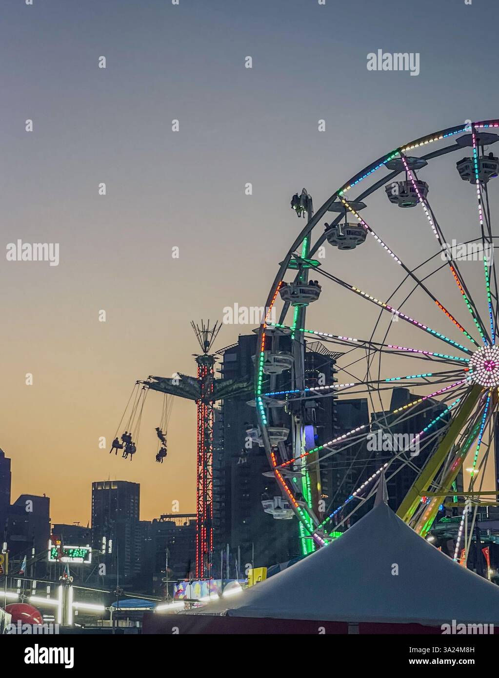 Ein beleuchtetes Riesenrad und eine drehende Schaukelfahrt vor der Skyline der Stadt in der Abenddämmerung. Die Szenerie des Vergnügungsparks besticht durch leuchtende Lichter und ein Gebäude mit Silhouette Stockfoto