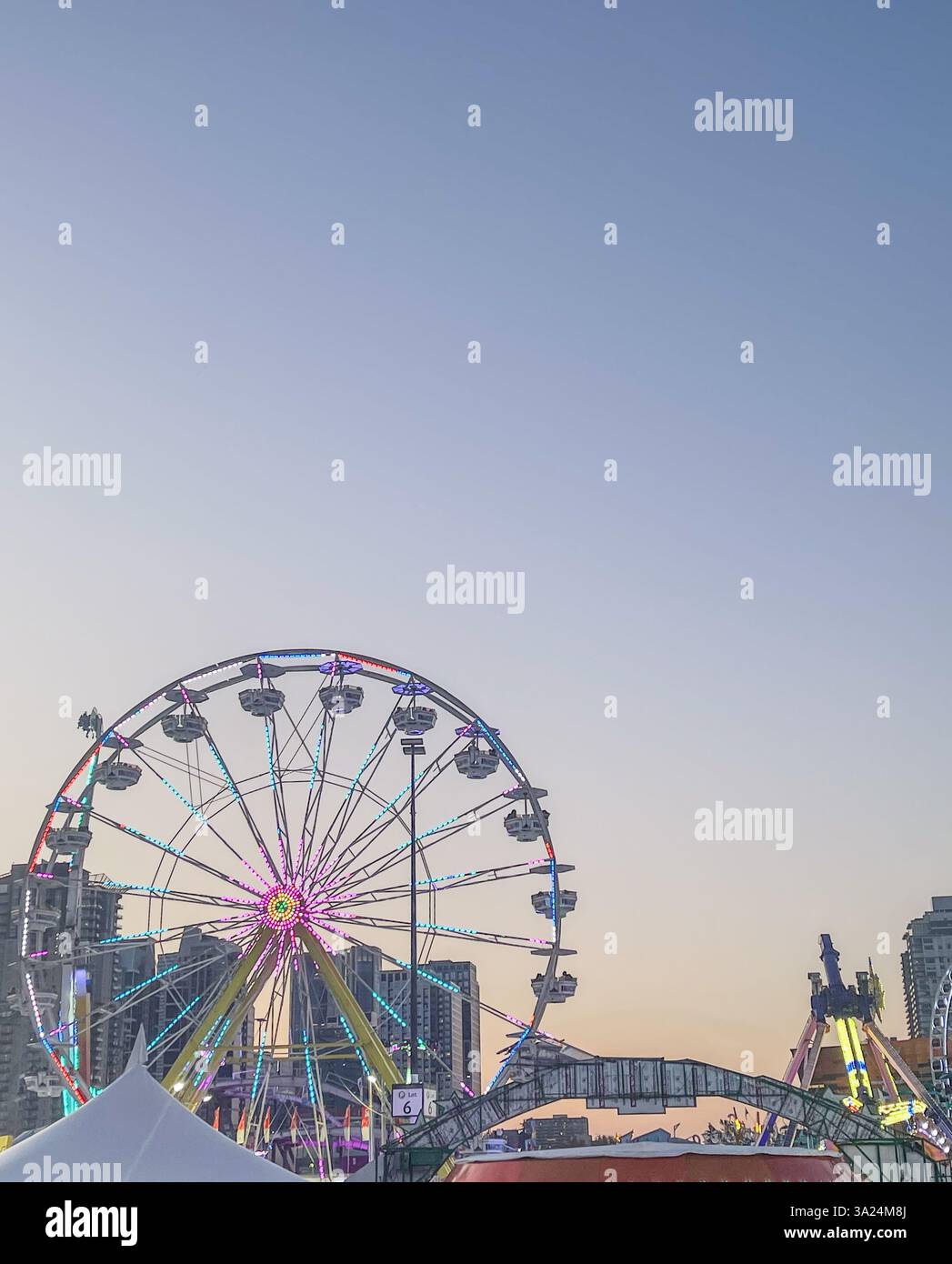 Ein großes Riesenrad mit hellen Lichtern vor einem weichen Dämmerhimmel in einem städtischen Vergnügungspark. Das Stadtbild im Hintergrund steht im Kontrast zum Th Stockfoto