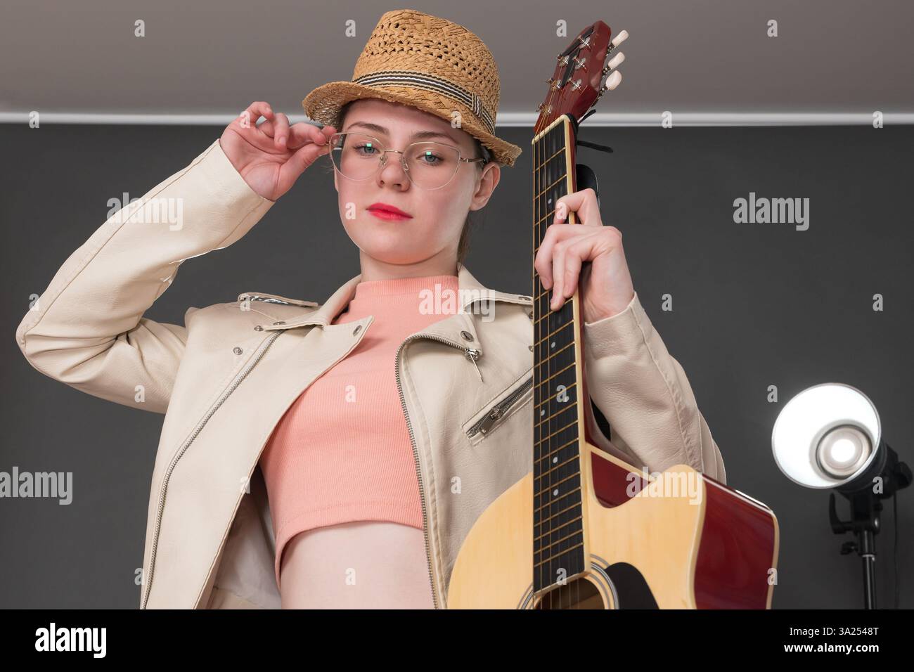 Porträt einer jungen Gitarristin mit Akustikgitarre in einer Hand, passt ihre Brille mit der anderen Hand an und blickt selbstbewusst in die Kamera. Musi Stockfoto