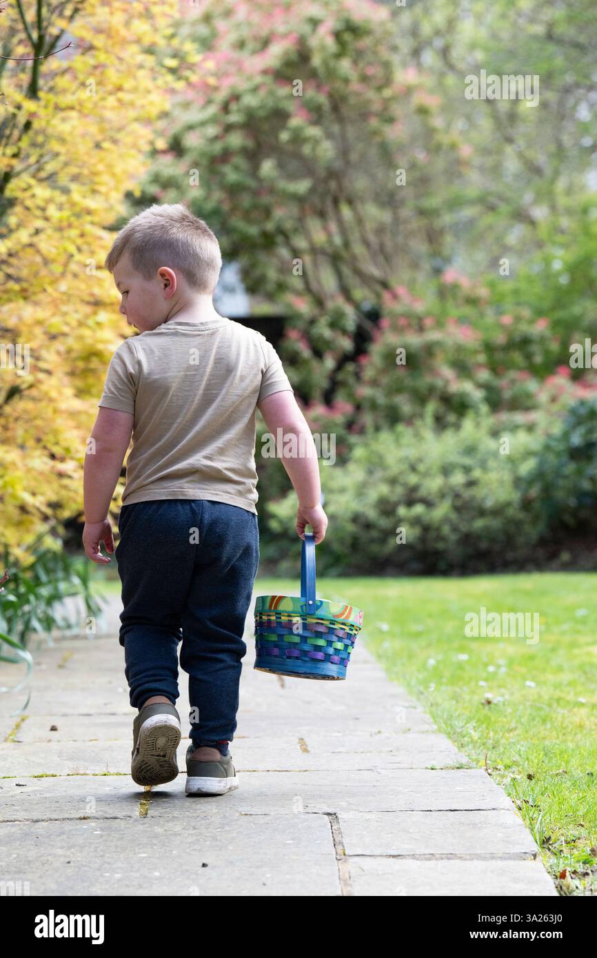 Rückansicht eines Jungen, der auf einem Gartenweg läuft und einen Korb mit Ostereiern trägt. Stockfoto