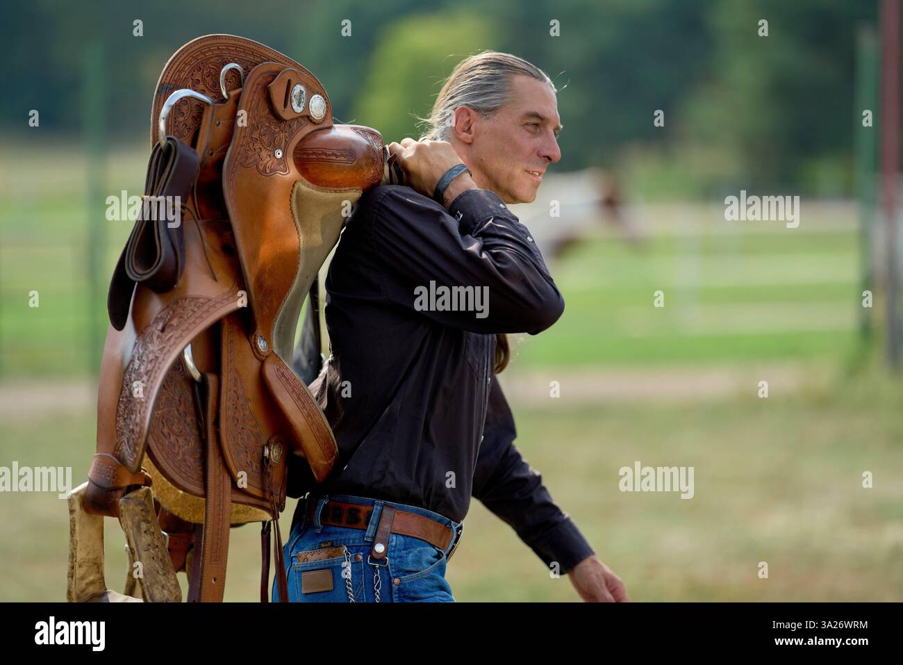Ein Mann in einem schwarzen Hemd trägt draußen einen braunen Sattel über der Schulter. Brandenburg, Deutschland Stockfoto