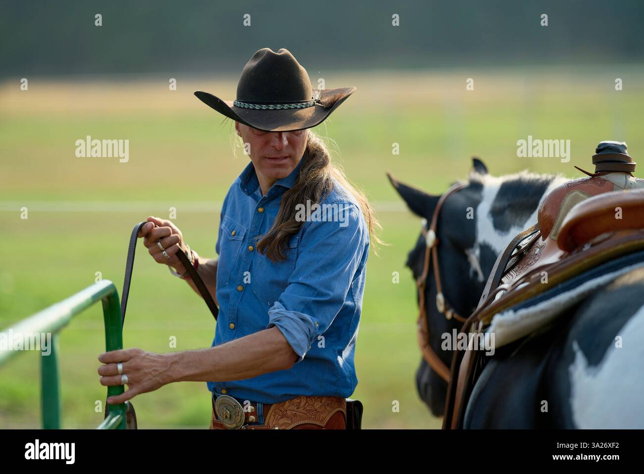 Cowboy in einem blauen Hemd hält ein Tor, mit einem gesattelten Pferd neben ihm auf einer Ranch. Brandenburg, Deutschland Stockfoto