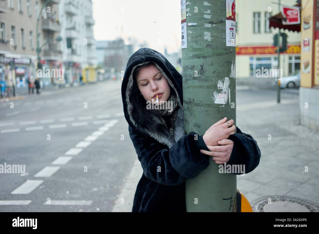 Eine Frau mit schwarzem Mantel und Kapuze steht auf einer Stadtstraße, hält einen Stab und raucht. Berlin, Deutschland Stockfoto
