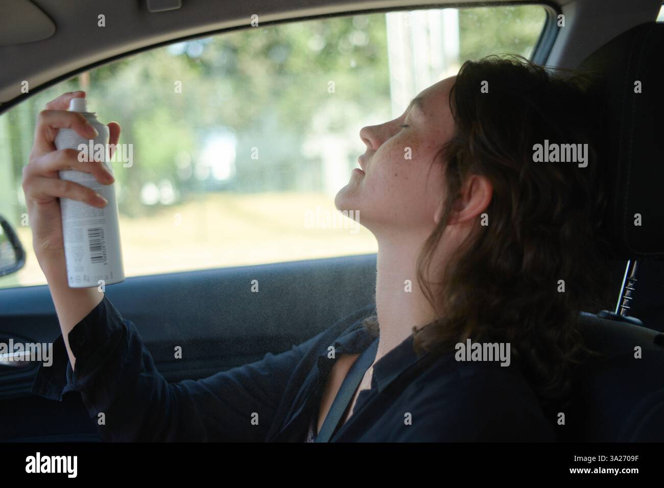 Frau, die Wasserspray in heißem Auto mit geschlossenen Augen und entspanntem Ausdruck an sonnigem Tag aufträgt. Stockfoto