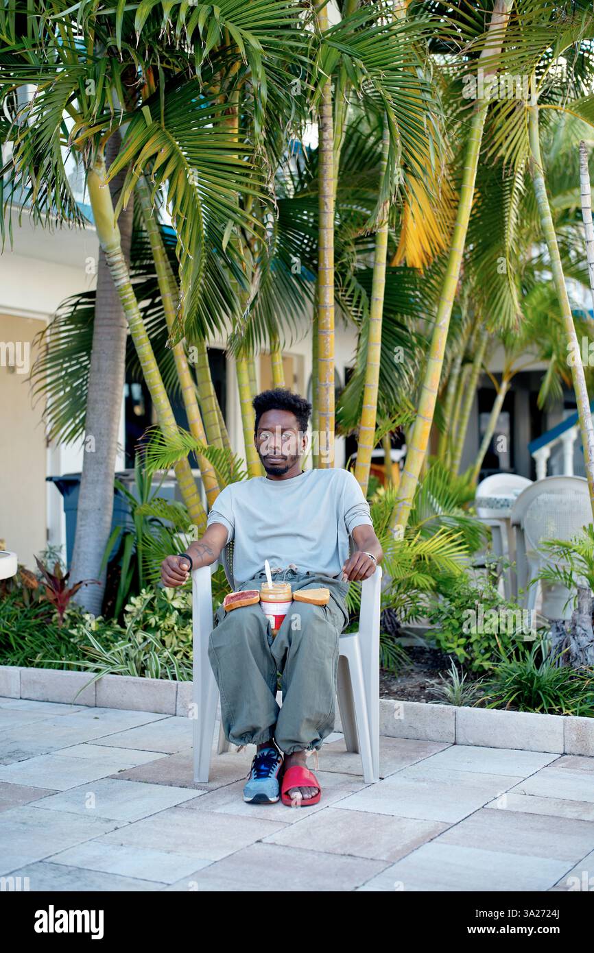 Mann sitzt draußen auf einem Stuhl, umgeben von Palmen und bereitet ein Erdnussbutter-Sandwich vor. Miami, Florida Stockfoto