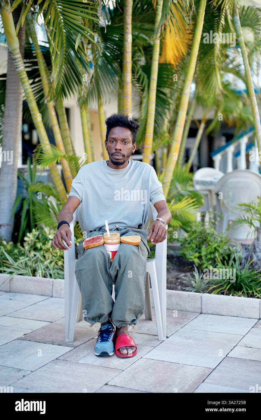 Mann sitzt draußen auf einem Stuhl, umgeben von Palmen und bereitet ein Erdnussbutter-Sandwich vor. Miami, Florida Stockfoto