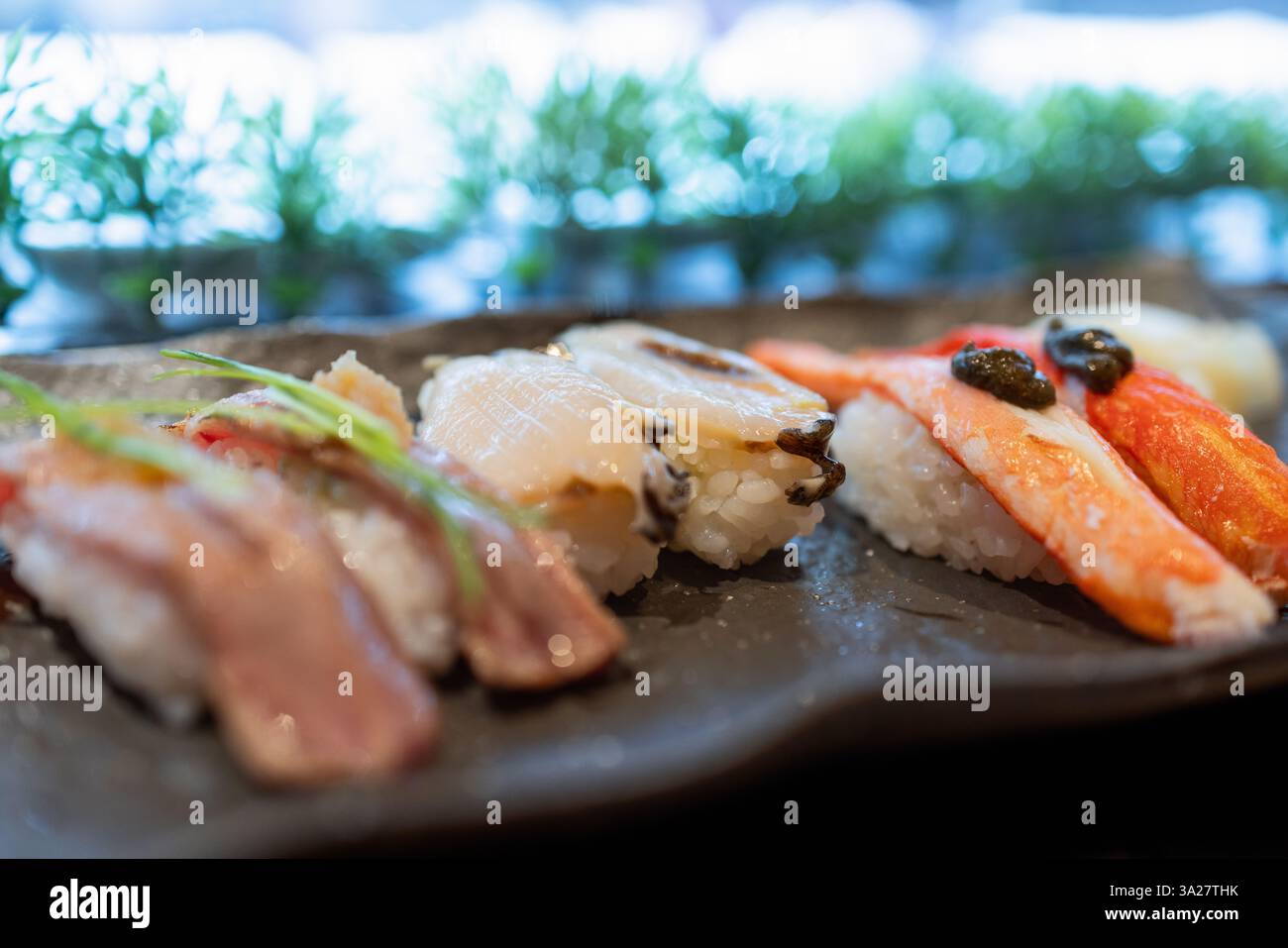 Verschiedenes Sushi mit flammengeröstetem Thunfisch, frischer Jakobsmuschel und Krabben-Nigiri auf einem dunklen Keramikteller, das die Essenz des japanischen Feinschmeckerdinners einfängt. Stockfoto