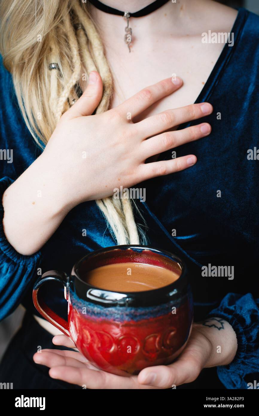 Blonde Frau mit Dreadlocks, die Kakaobecher und Hand am Herzen hält, mit Halskette mit Schlangenanhänger Stockfoto