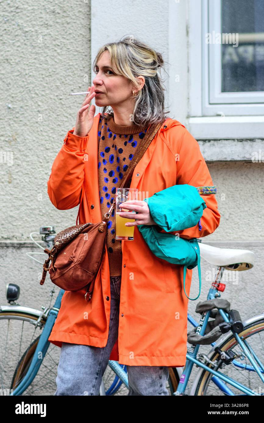 Junge Frau mit langen blonden Haaren, die eine leuchtend orange Jacke trägt und auf der Straße steht und raucht - Tours, Indre-et-Loire (37), Frankreich. Stockfoto