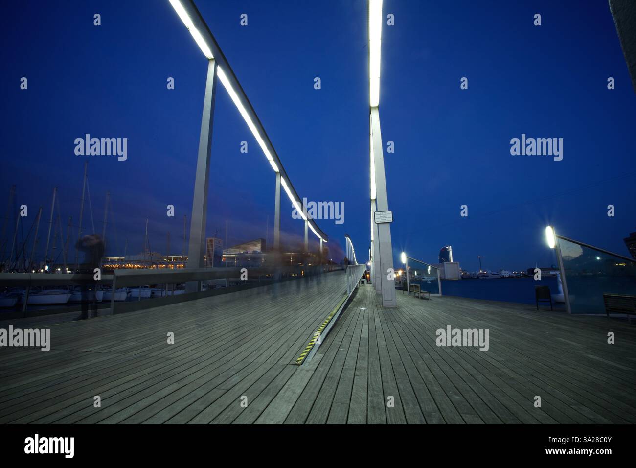 Ein moderner Holzsteg in der Abenddämmerung, beleuchtet von hellen Deckenleuchten. Die Szene zeigt einen Yachthafen mit Booten im Hintergrund, was eine ruhige Atmosphäre schafft Stockfoto
